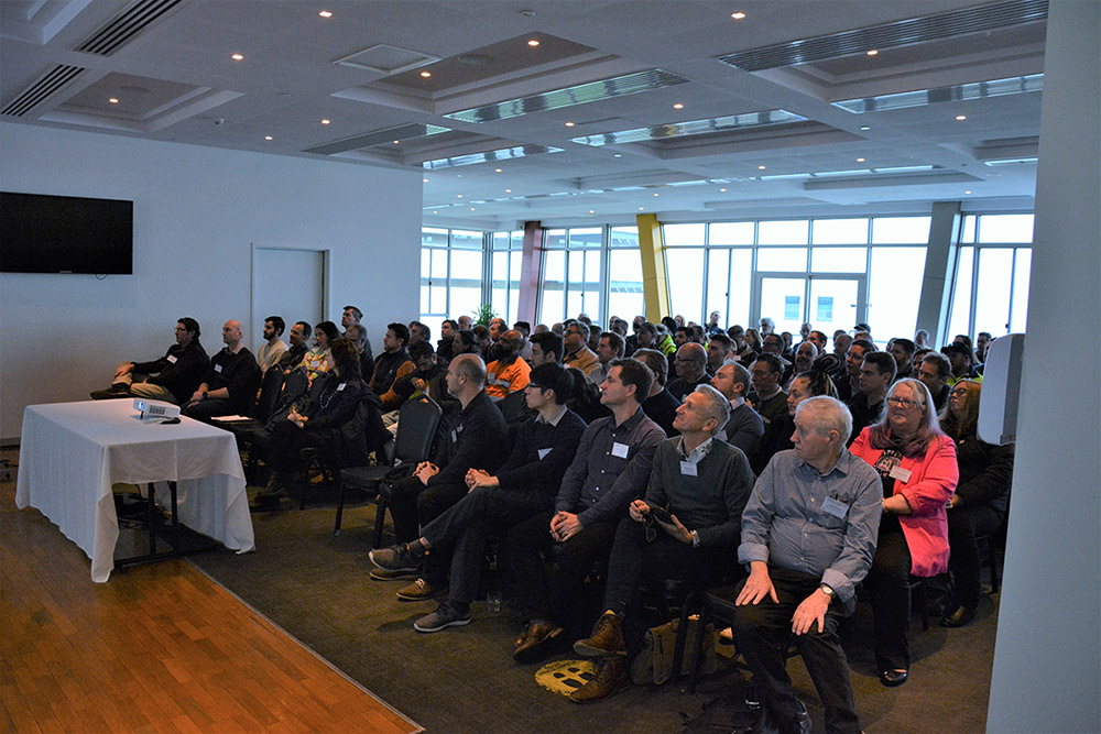 An audience of about 100 people sit and listen to a speech.