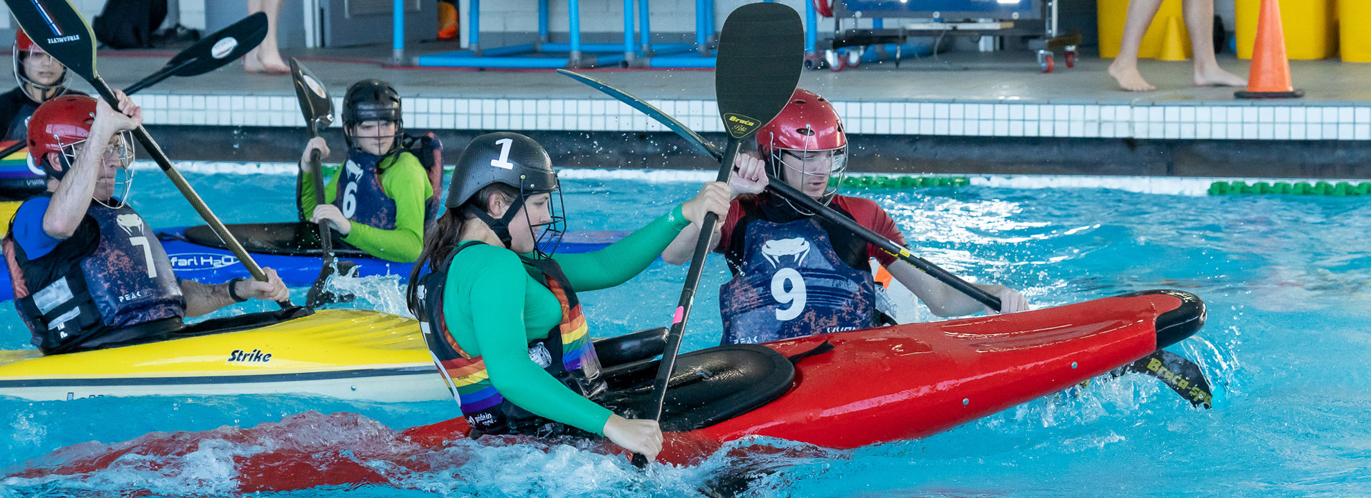 Action shot of young people in a pool using kayaks to play a game of polo. Their paddles are in the air and the water in splashing.