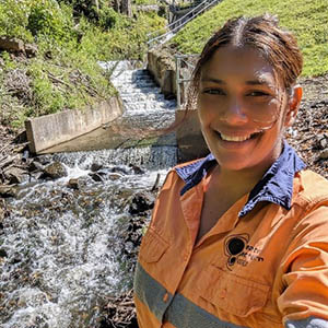 Adlin is taking a selfie near some running water and greenery in the background. She is wearing a hi-viz shirt.