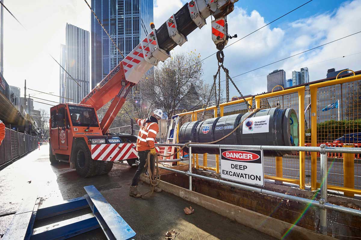 A lagre orange crane lowers a section of sewer pipe into the excavation hole.