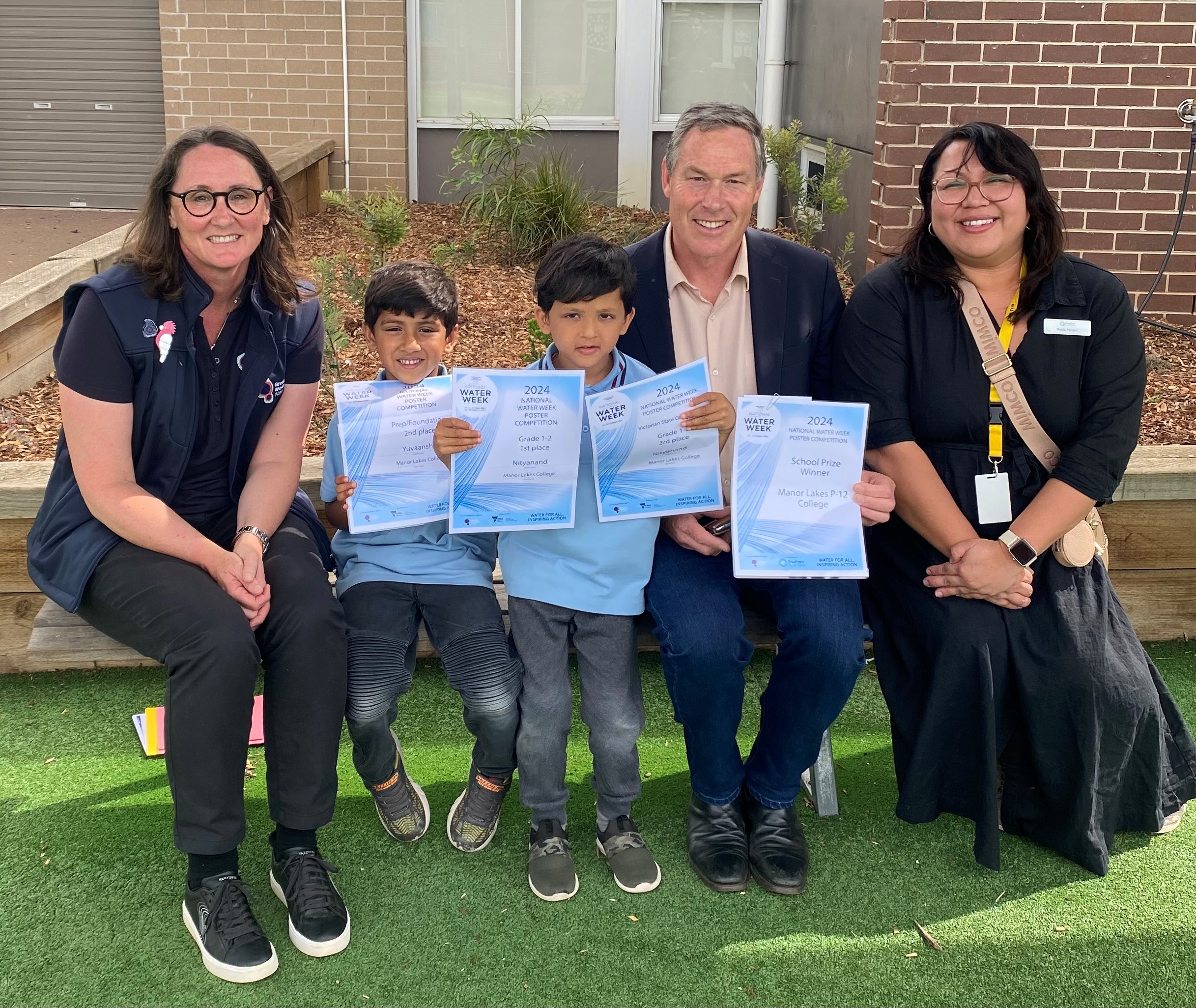 Three adults and two children, smiling with their National Water Week Poster Competition certificates, seated outside on a bench. The children are wearing blue school uniforms. 