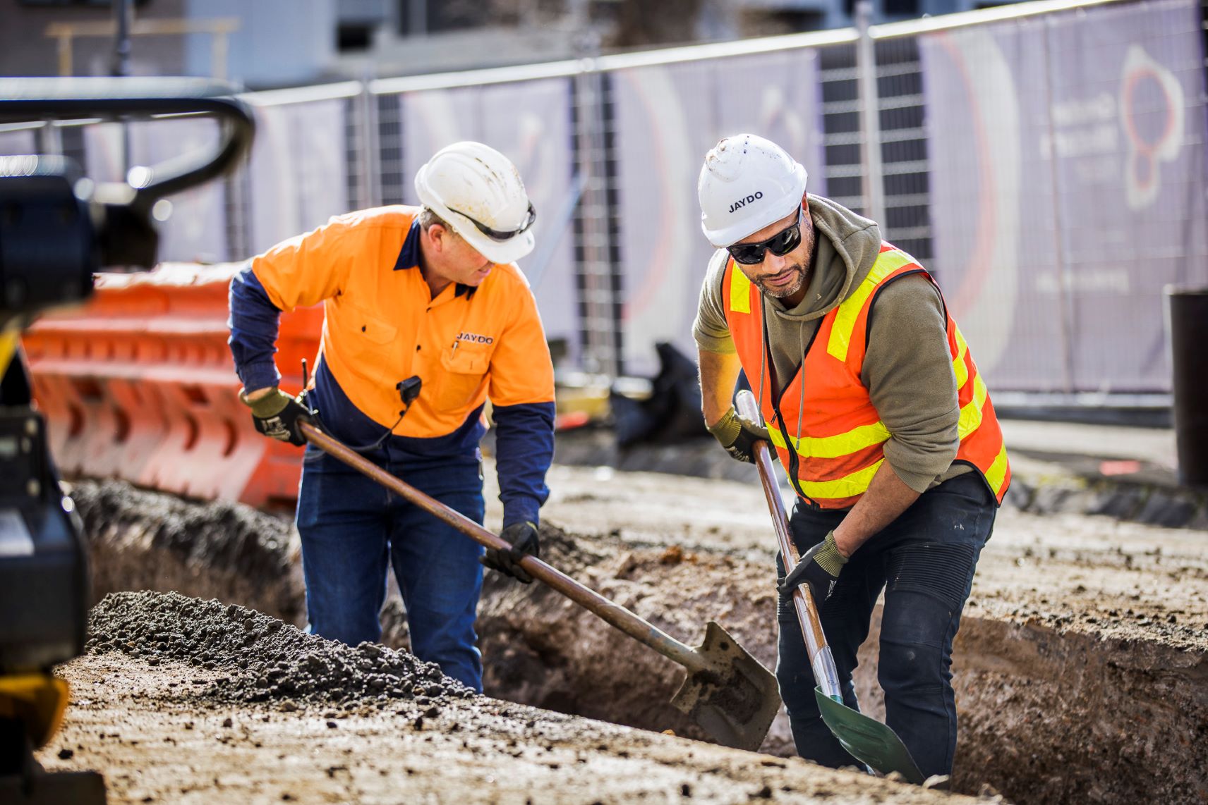 Two men on a construction site dressed in PPE holding shovels 