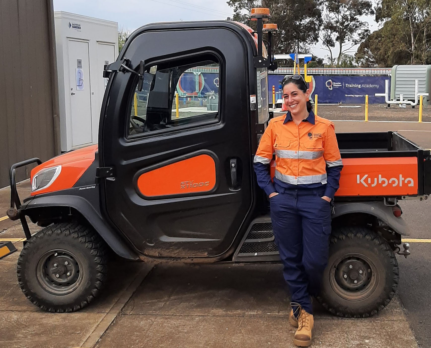 A woman wearing a Greater Western Water branded uniform standing casually in front of a small pick-up truck.