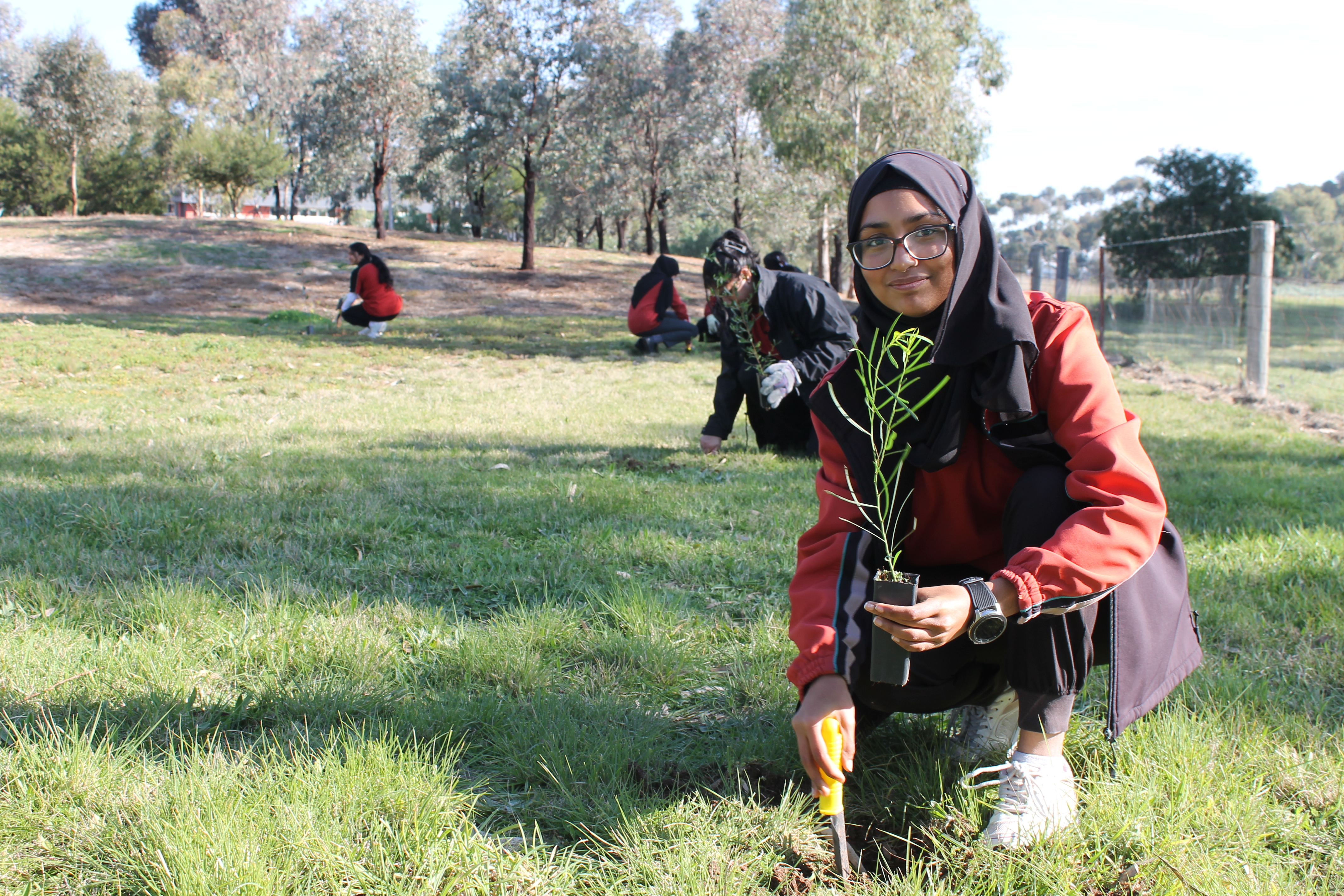 A student in a red and black uniform in a grassy area about to plant a young tree in the ground, with other students in the background