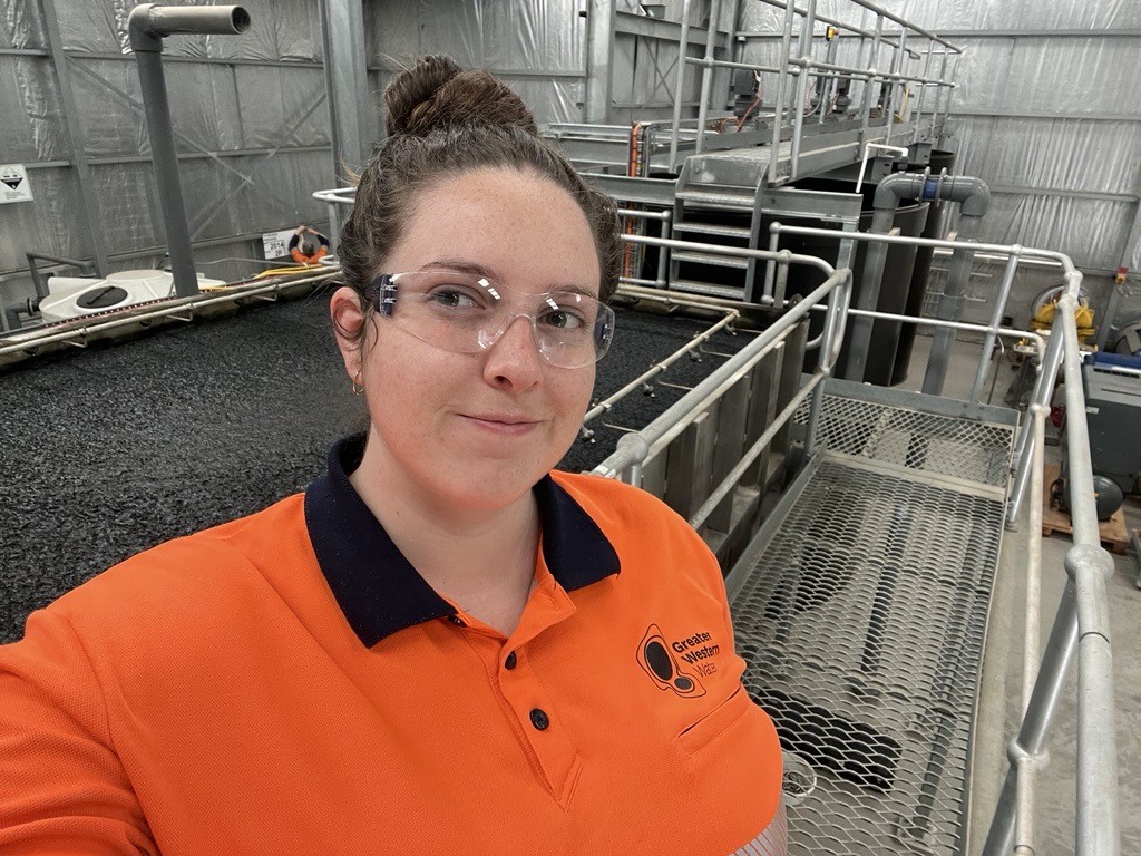 Jacki is in an orange work shirt with a GWW logo, wearing safety glasses, standing in an industrial setting with metal structures and equipment in the background.