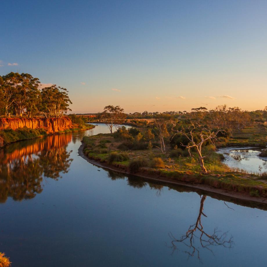 The Werribee River, photograph taken from the bank so showing the deep blue colour of the water and the sun highlighting the red rocks making up the bank of the river.