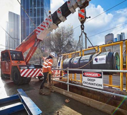 A lagre orange crane lowers a section of sewer pipe into the excavation hole.